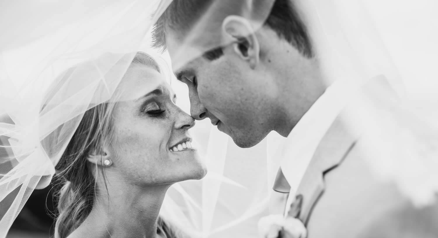 black and white photo of smiling woman and man in a tuxedo with noses touches and a wedding veil covering both of their heads.