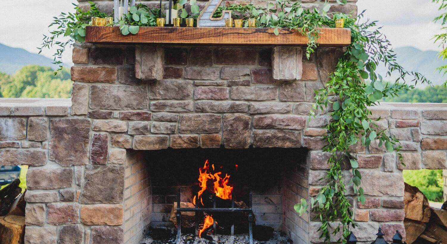 outdoor stone fireplace with tall stone chimney and wooden mantle, with an orange glowing fire, and mountains in the background.