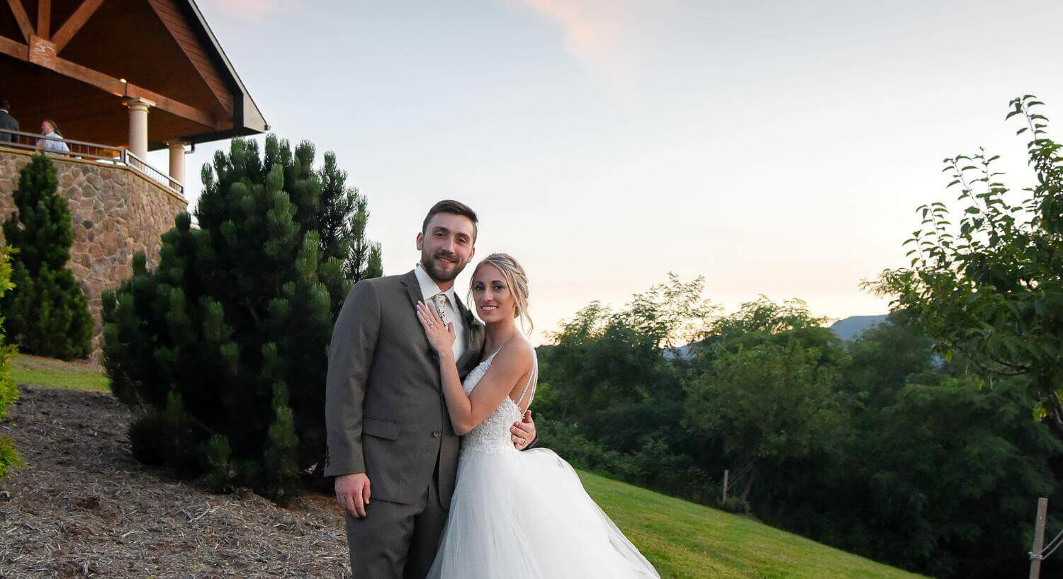 embracing wedding couple in a grey suite, white wedding dress, on green grass with a sone and wood building in the background, with green trees and a blue-pink sky in the background.