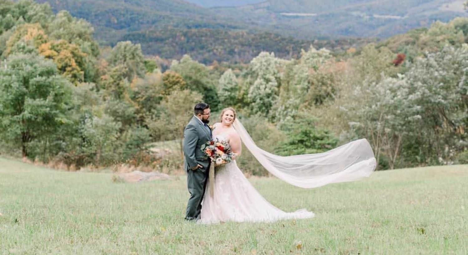 wedding couple in a grey suit and white dress with wedding veil blowing in the wind, holding a bouquet of colorful flowers, standing in green grass, with trees and mountains in the background