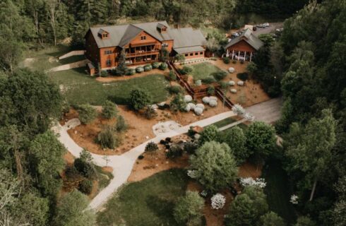 Aerial photo of wood buildings, surrounded by green grass, trees, driveways, sidewalks, and a large pond.