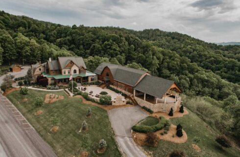 Aerial photo of stone and wood buildings, surrounded by green grass, trees on hills, and paved driveways.