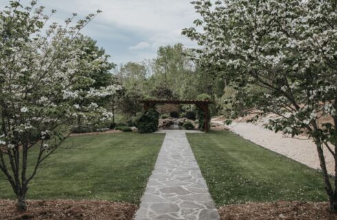 Outdoor stone walkway in between green grass, trees with white flowers, leading to a wood pergola, with trees and bushes in the background.
