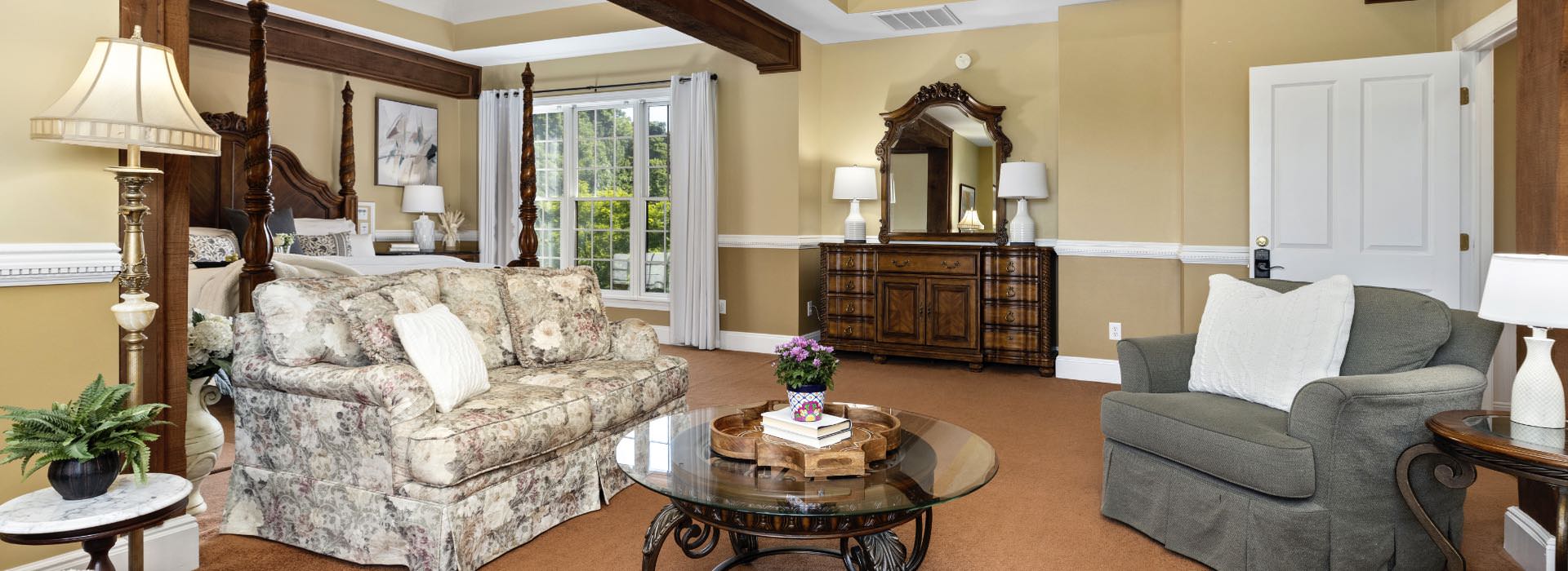 Large bedroom suite with light tan walls, white trim, carpeting, four-poster bed, wooden dresser, and sitting area