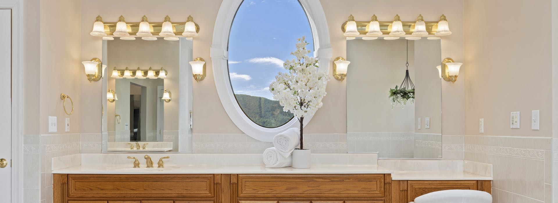 Large bathroom with light colored walls, wooden vanity with white countertop, brass fixtures, large oval mirror
