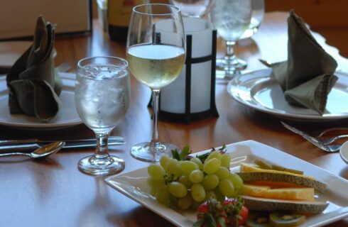brown table with a plate of fruit, a glass of wine, red roses, and a wine list next to a bottle of wine, with a window and greenery behind the table.