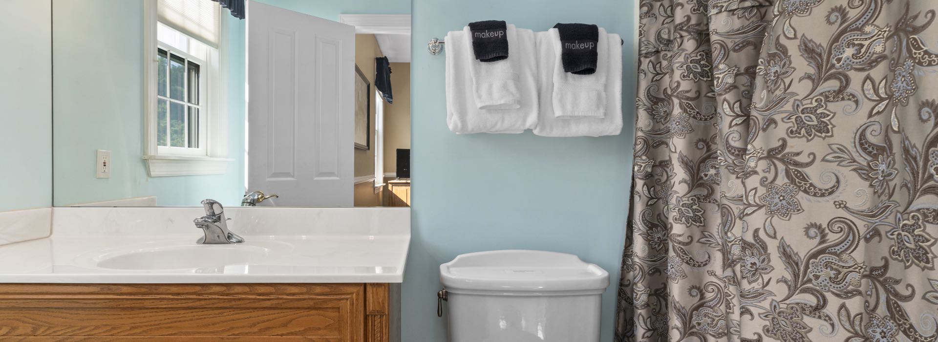 Bathroom with light blue walls, white toilet, wooden vanity with white counter top, and paisley shower curtain