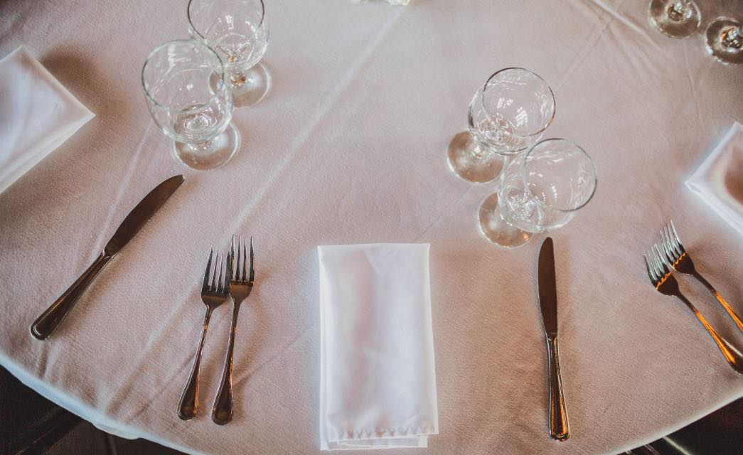 Round dining table with white tablecloth and napkins, silver forks and knives, and glass stemware.