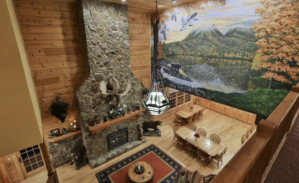Dining room from upper floor of wood floor, wood dining tables and chairs, floor to ceiling stone fireplace, and red and black rug with circular table, along with a floor to ceiling mural of mountains, trees, grass, and pond.