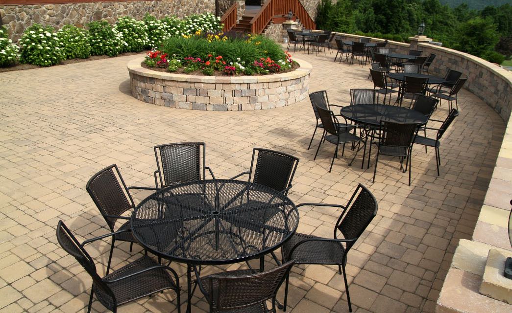Outside patio with stone floor and partial walls, black wrought iron circular tables and chairs, and circular stone flower bed with flowers and greenery, surrounded by trees and bushes.