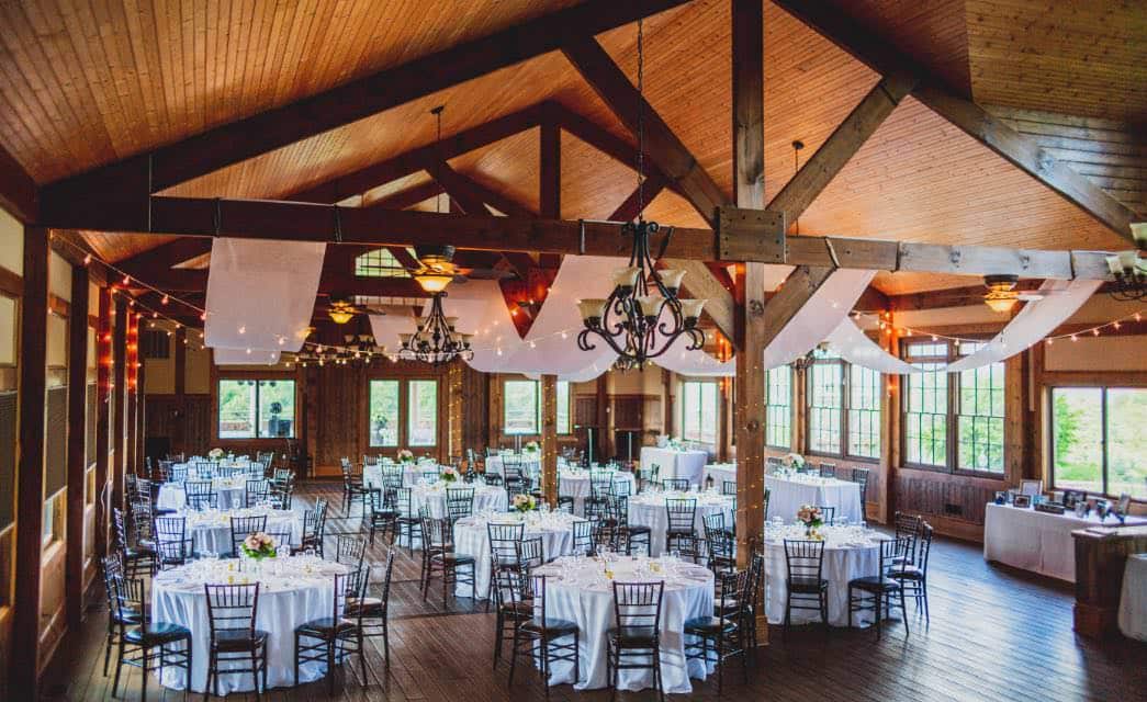 Venue with large wooden ceiling and wood beams, wood floors, circular tables with white tablecloths and black chairs, with lots of surrounding windows, and lights and curtains draped from the beams.