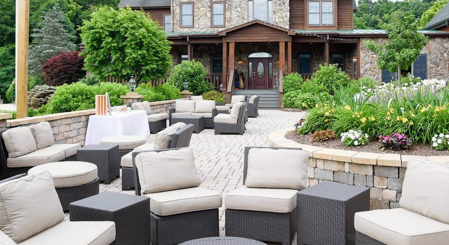 outdoor sitting area on a stone patio with dark brown wicker furniture and beige cushions, stone and wood house in the background with stone flower box and green trees and bushes.