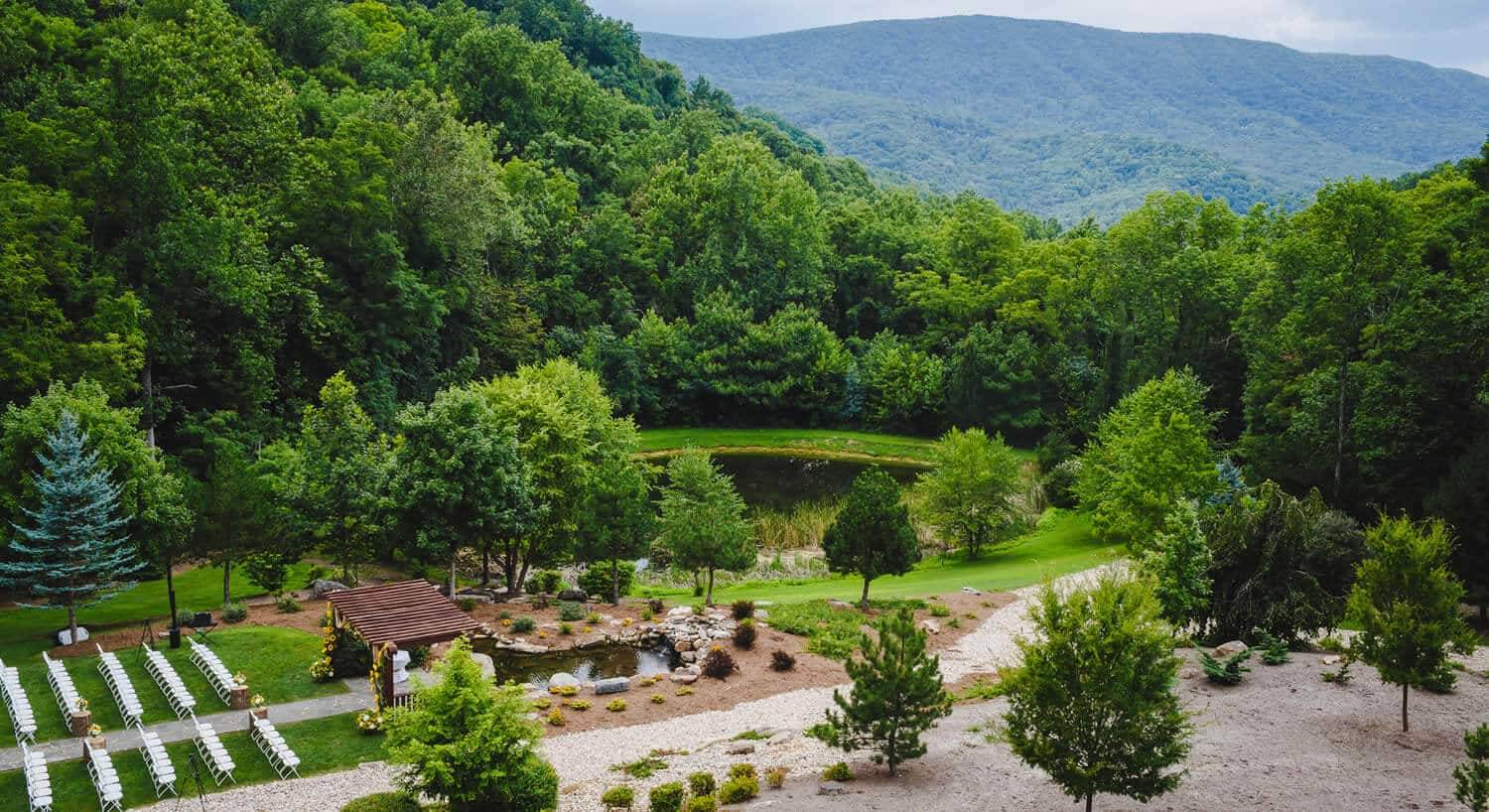 outdoor wedding venue with dark brown wooden pergola, white chairs, 2 ponds, green grass and trees and blue-green mountains in the background.