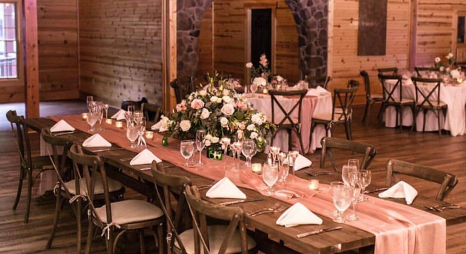 large room with wooden floors and walls, and stone archway set up for a wedding reception with wood tables and chairs, beige cushions, pink fabric, bouquets of flowers, and lights hanging from the wooden beams