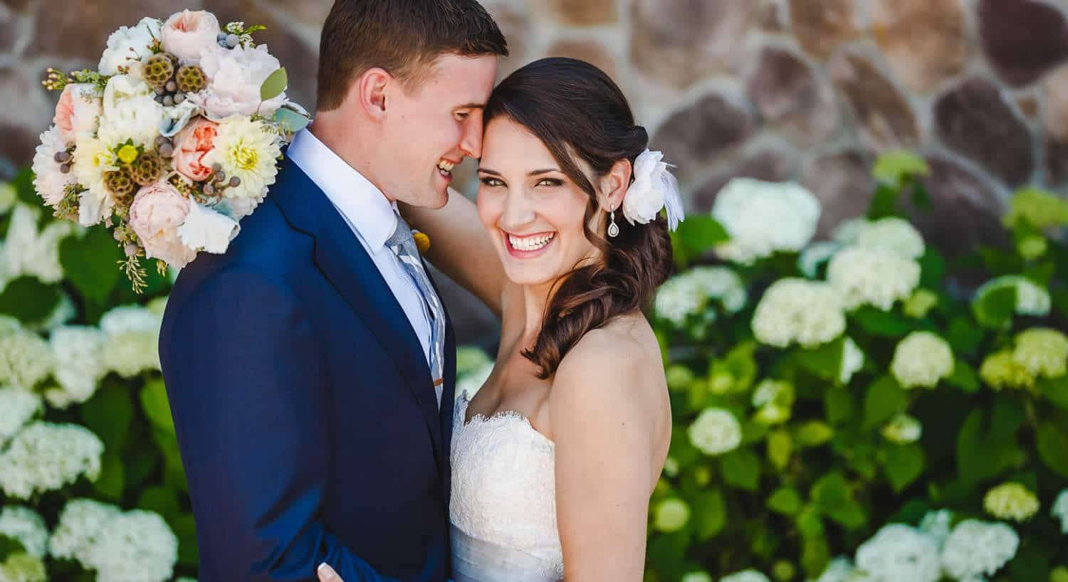 smiling, embracing wedding couple with a man in a blue suit, woman in a white dress with white flower in her hair, floral bouquet, in front of green bushes with white flowers and a stone wall