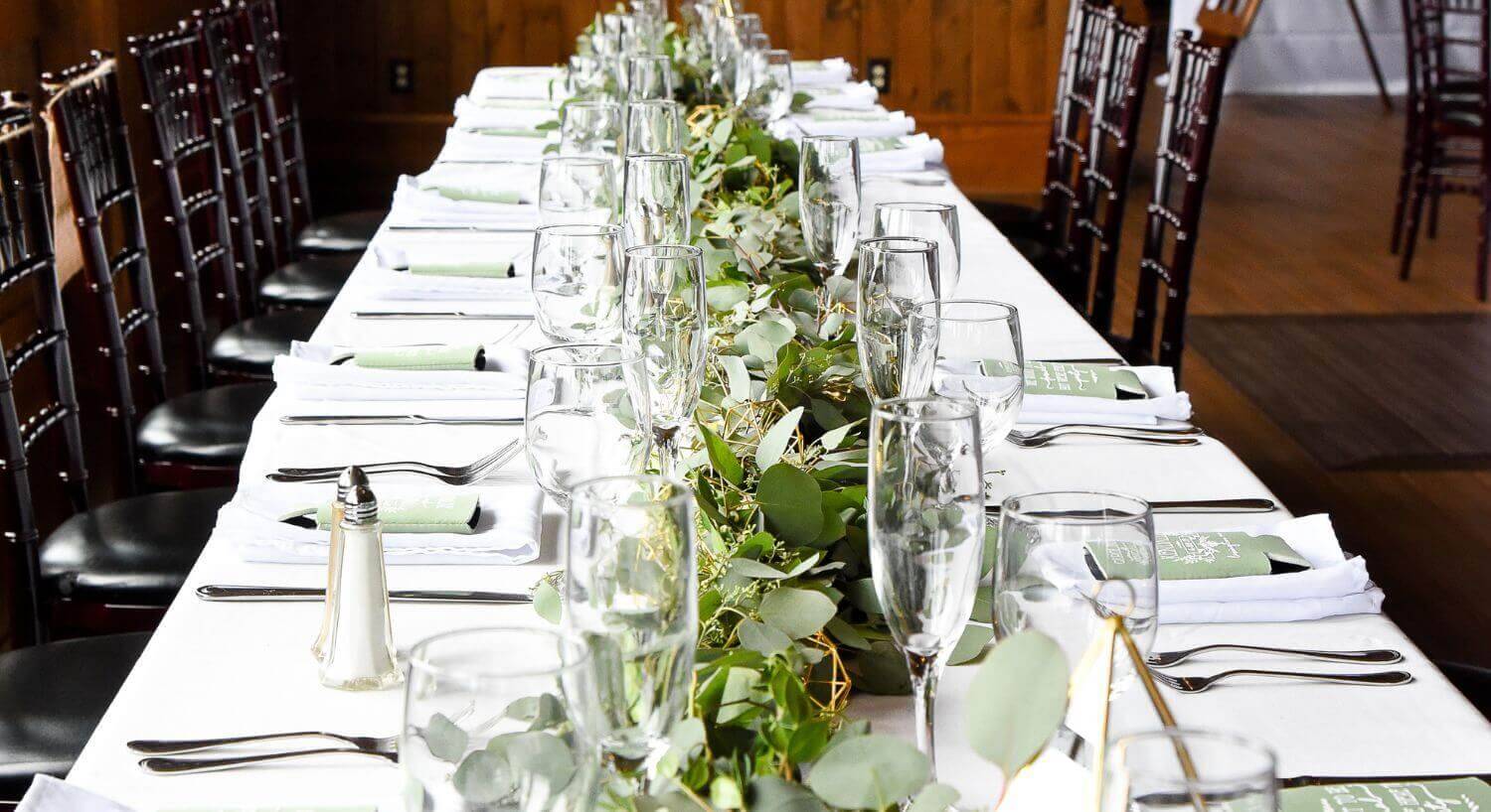long white cloth covered table with greenery in the middle, champagne glasses, silverware, and dark wooden chairs, set up for a reception in a room with wood floors, walls, and accents